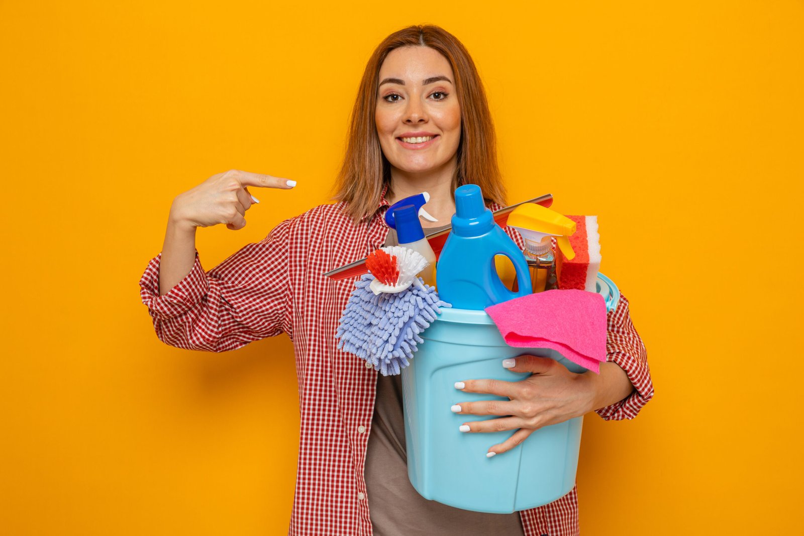 happy young cleaning woman in plaid shirt holding bucket with cleaning tools looking at camera smiling cheerfully pointing with index finger at herself standing over orange background